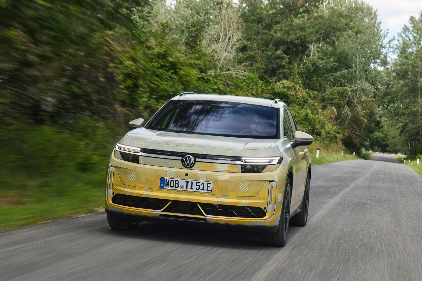 Jasnożółty samochód elektryczny marki Volkswagen jedzie po asfaltowej drodze otoczonej zielenią i drzewami.