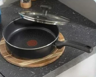 Większa patelnia Tefal Czarna patelnia z pokrywką leżąca na okrągłej drewnianej desce na kuchennym blacie.