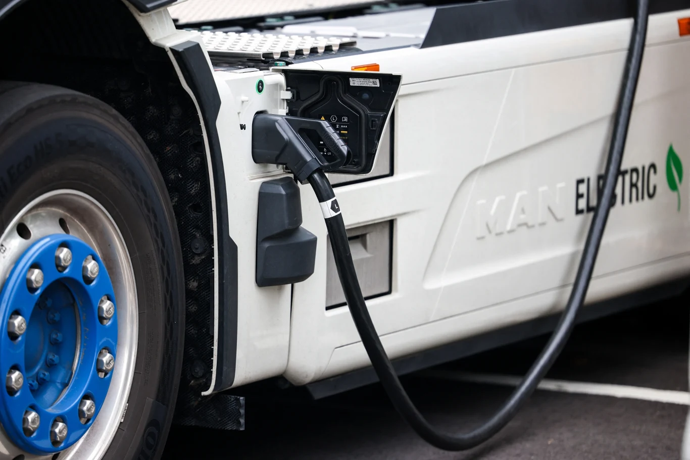 Elektryczna ciężarówka podłączona do stacji ładowania, widoczne zbliżenie na przewód oraz port ładowania umieszczony przy bocznej części pojazdu.