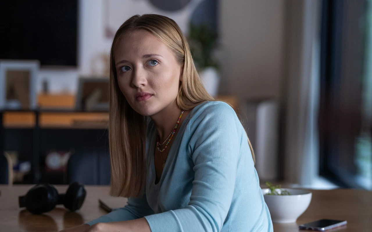 Karolina Matej na planie "Zdrady" Młoda kobieta z blond włosami w jasnoniebieskim swetrze siedzi przy stole, obok leży miska z jedzeniem, słuchawki oraz telefon.