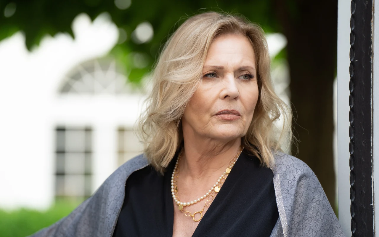 Grażyna Szapołowska na planie "Zdrady" Kobieta w średnim wieku z blond włosami, ubrana elegancko w ciemny strój i perłowy naszyjnik, stoi na tle rozmytej architektury i zieleni.