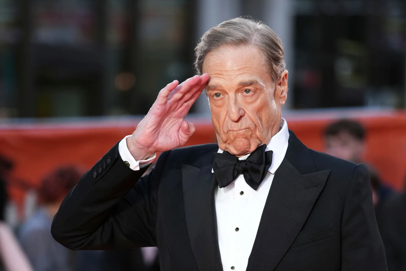 John Goodman John Goodman w smokingu z muchą salutuje, stojąc na tle rozmytego tłumu.