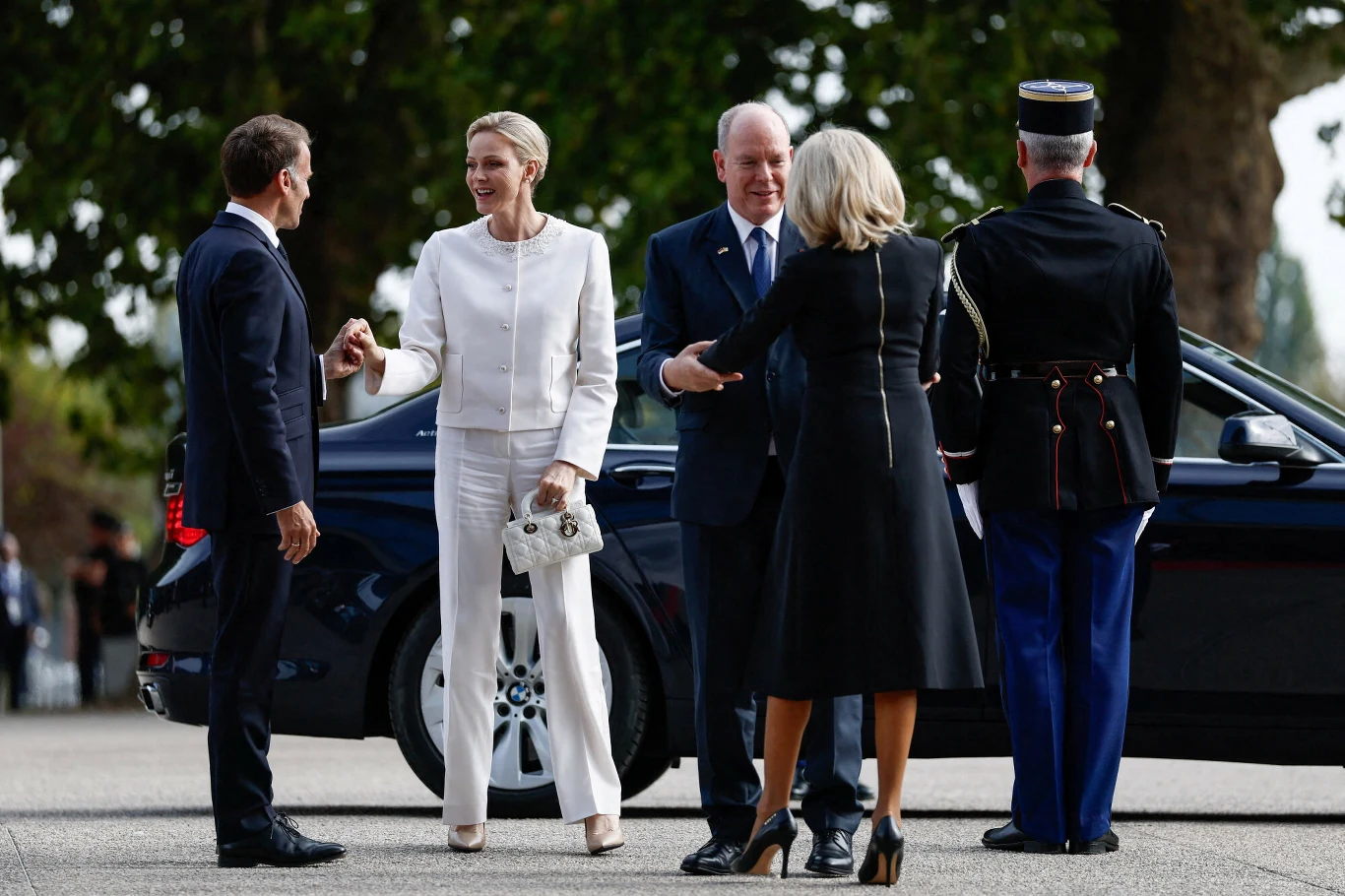 Grupa elegancko ubranych osób wita się przy czarnym samochodzie, w tle funkcjonariusz w mundurze i zieleń drzew.