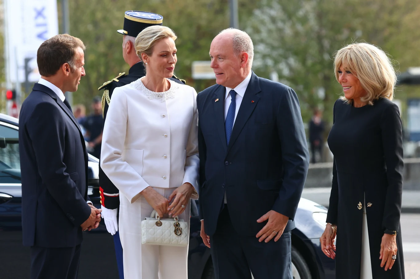 Cztery elegancko ubrane osoby prowadzą rozmowę na zewnątrz, w tle rozmyta sylwetka samochodu, za nimi stoi mundurowy funkcjonariusz.