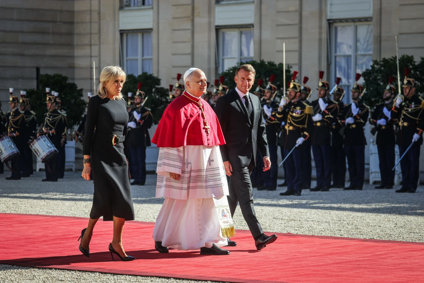 Trzy osoby w eleganckich strojach idą czerwonym dywanem, w tle szpaler żołnierzy w galowych mundurach oraz historyczny budynek.