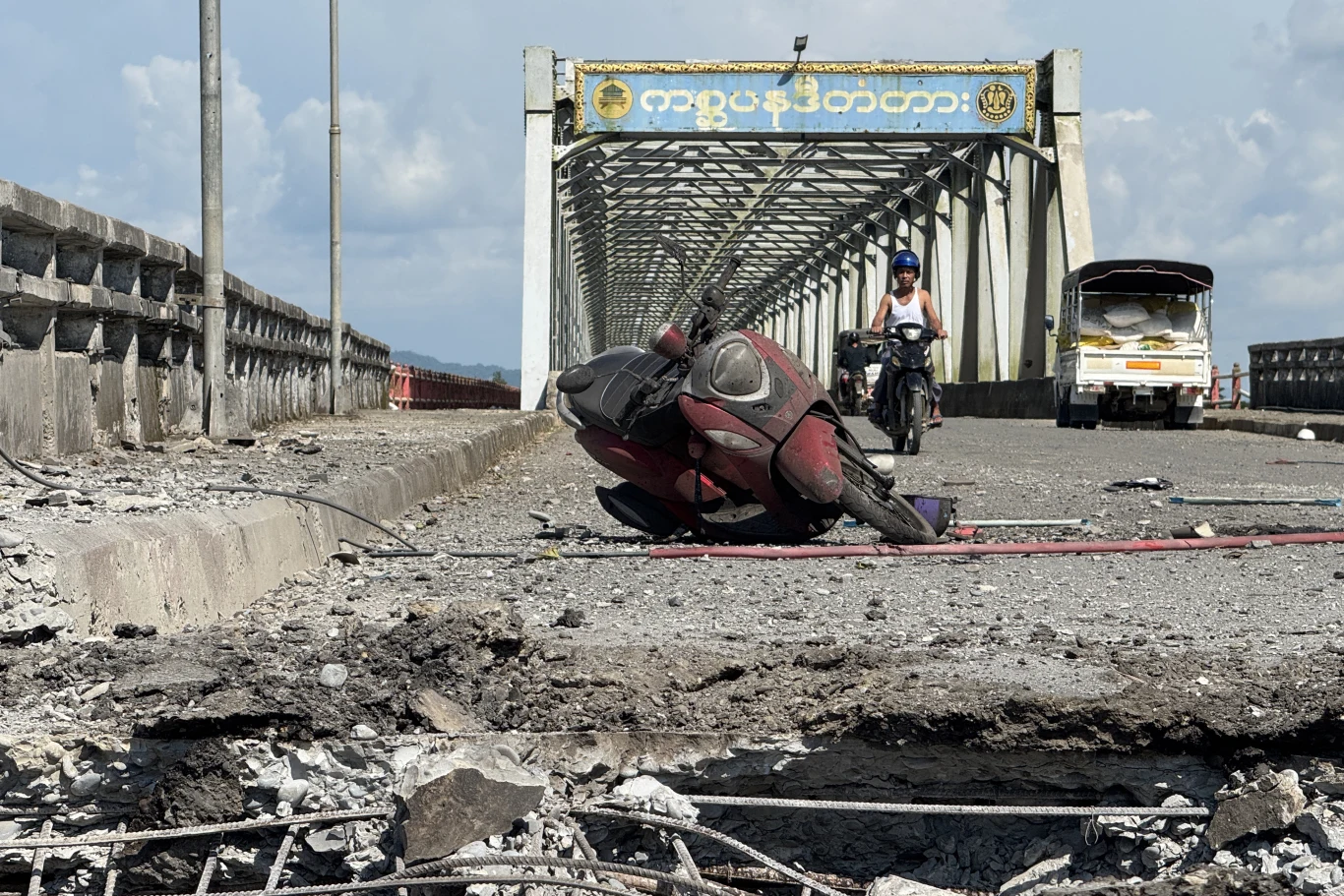 Skutki ataku junty wojskowej na bazar w Kyauktaw na pograniczu z Indiami Uszkodzony most z dużą wyrwą w jezdni, przewrócony skuter leżący na zniszczonej nawierzchni oraz mężczyzna jadący rowerem, a w tle biały pojazd typu tuk-tuk pod żółtym napisem na metalowej konstrukcji mostu.