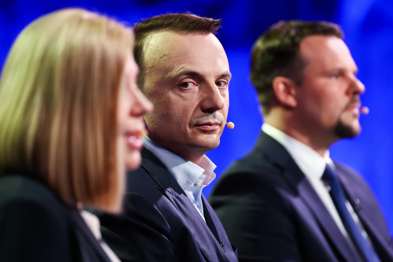 Trójka osób ubranych formalnie siedzi obok siebie podczas wydarzenia, wszystkie mają niewielkie mikrofony przy ustach, a środkowa postać patrzy prosto w kamerę.
