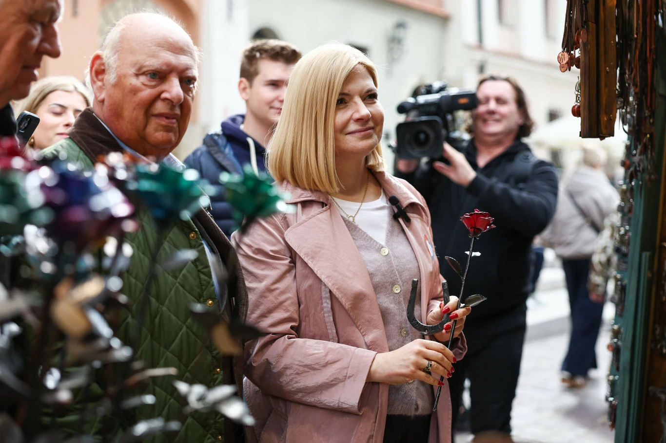 Kobieta w jasnym płaszczu trzymająca metalową różę, stojąca obok starszego mężczyzny na targu rękodzieła, w otoczeniu innych osób i fotografa z kamerą.