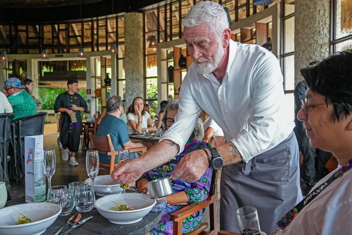 Brodaty mężczyzna w białej koszuli nalewa sos do talerza siedzącej osoby w eleganckiej restauracji, gdzie inni goście rozmawiają przy stole.