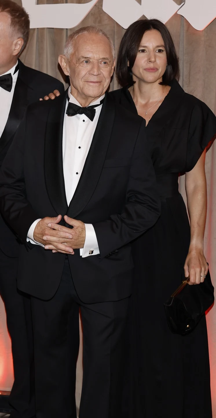 Marek Kondrat i Antonina Turnau Elegancko ubrany mężczyzna w smokingu stoi obok kobiety w długiej czarnej sukni, która trzyma małą torebkę w dłoni; oboje prezentują się oficjalnie i pozują do zdjęcia na tle jasnej dekoracji.