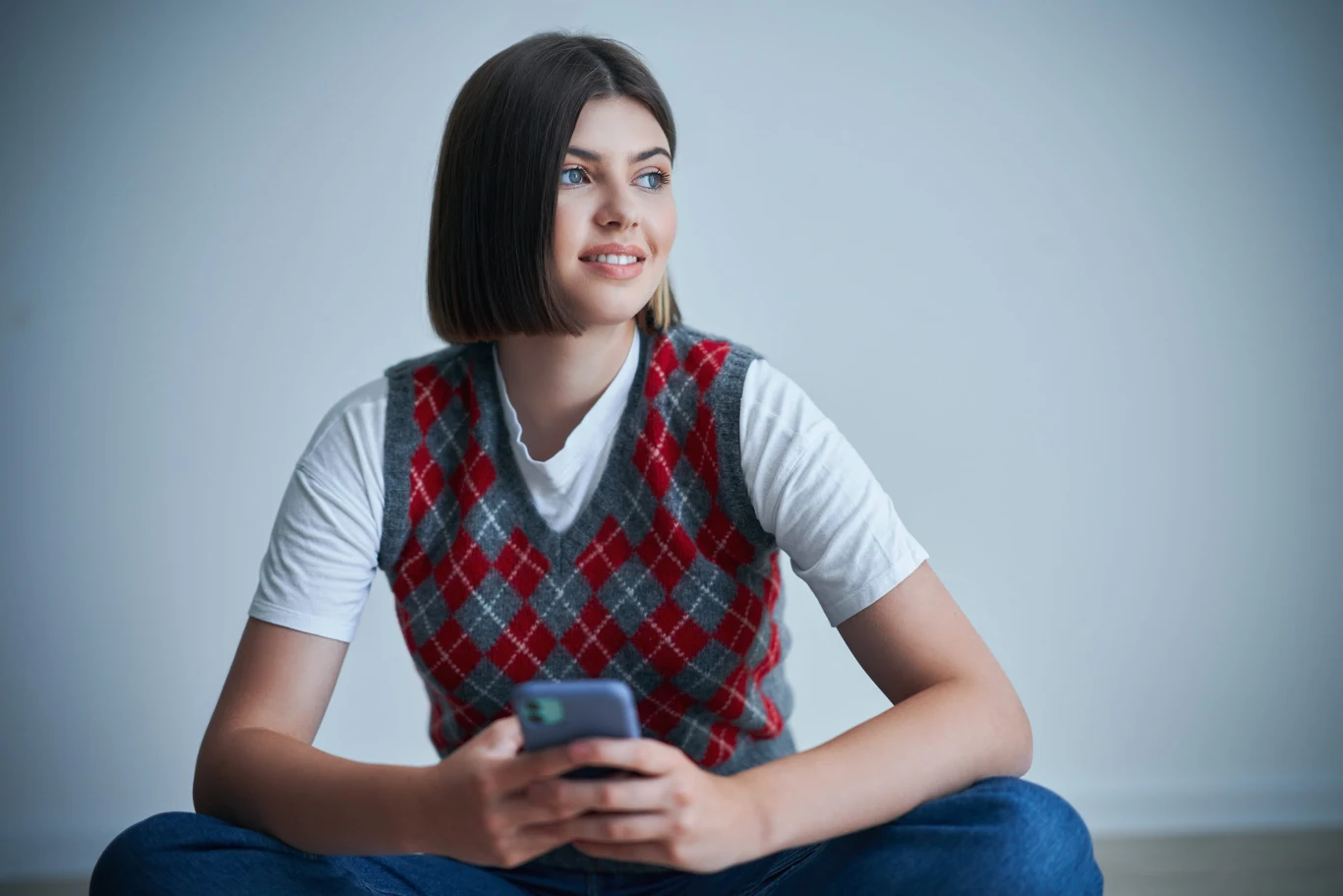 Młoda kobieta z krótkimi włosami, ubrana w biały t-shirt i sweter w czerwoną kratę, siedzi ze skrzyżowanymi nogami, trzymając smartfon i patrząc w bok z lekkim uśmiechem.
