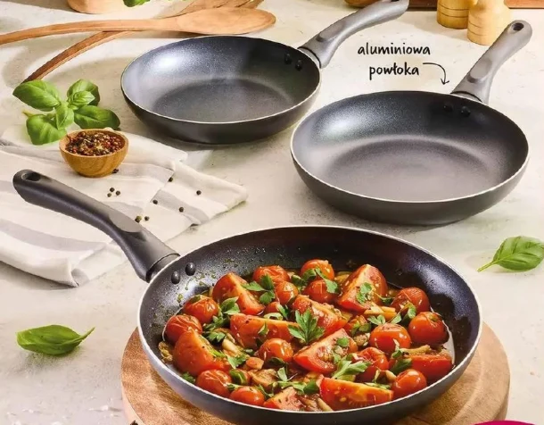 Zestaw trzech patelni Magdy Gessler za 79,90 zł Trzy patelnie z aluminiową powłoką, z których jedna wypełniona jest duszonymi pomidorami z ziołami, ustawione na jasnym blacie wśród świeżej bazylii i przypraw.