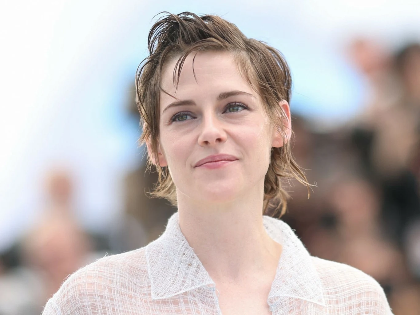 Kristen Stewart Uśmiechnięta kobieta z krótkimi, roztrzepanymi, jasnobrązowymi włosami ubrana w jasną, ażurową koszulę.