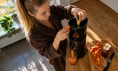 Kobieta wrzuca kawałki marchwi i jabłka do czarnej wyciskarki wolnoobrotowej, z której sok spływa do szklanki