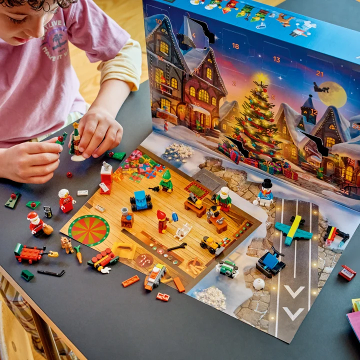 Dziecko układa małe klocki LEGO na rozłożonym kartonowym kalendarzu adwentowym z zimową, świąteczną scenerią i kolorową choinką, otoczone figurkami postaci oraz świątecznymi akcesoriami.