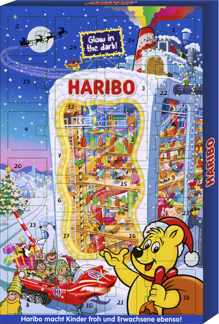 Kolorowa fabryka HARIBO pełna świątecznych dekoracji i postaci przygotowujących słodycze, na pierwszym planie żółty miś w czapce Mikołaja machający łapą, po lewej dzieci na saniach obsypanych żelkami, w tle zimowy krajobraz z saniami Mikołaja na niebie.