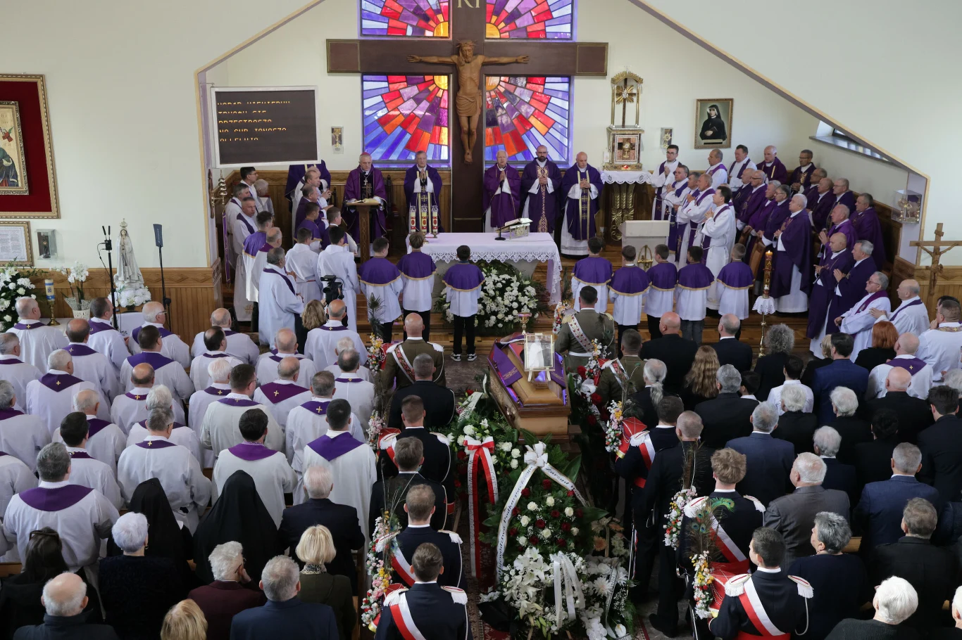 Uroczystość pogrzebowa w kościele, trumna otoczona kwiatami ustawiona przed ołtarzem, liczni duchowni w fioletowych szatach i zgromadzeni wierni, w tle duży krzyż i barwne witraże.
