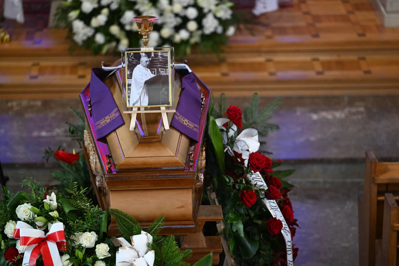 Trumna ozdobiona szarfami i kwiatami, na której stoi fotografia zmarłej osoby, otoczona wieńcami oraz bukietami białych i czerwonych róż.