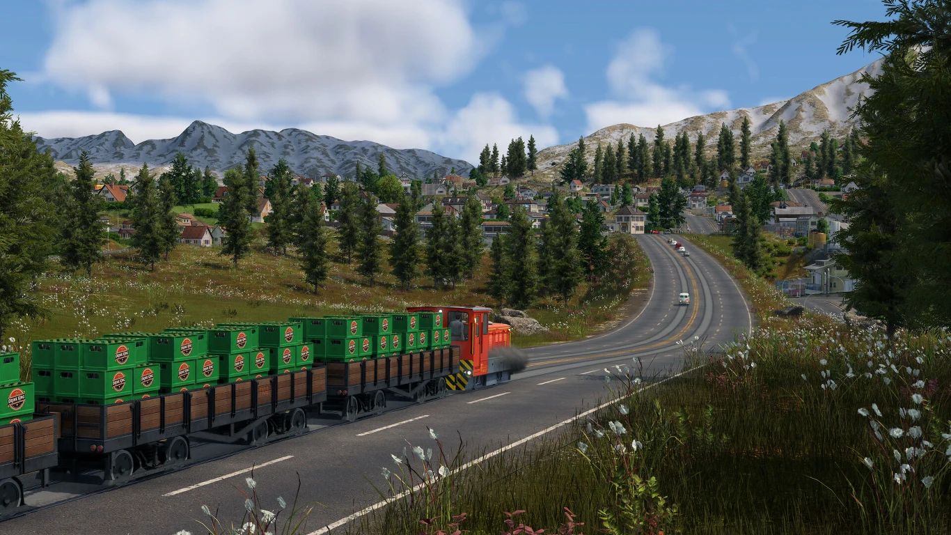 Transport Fever 3 Czerwony pociąg towarowy przewożący zielone kontenery porusza się wzdłuż asfaltowej drogi w górskiej okolicy, wśród drzew i w pobliżu zabudowań małego miasteczka.