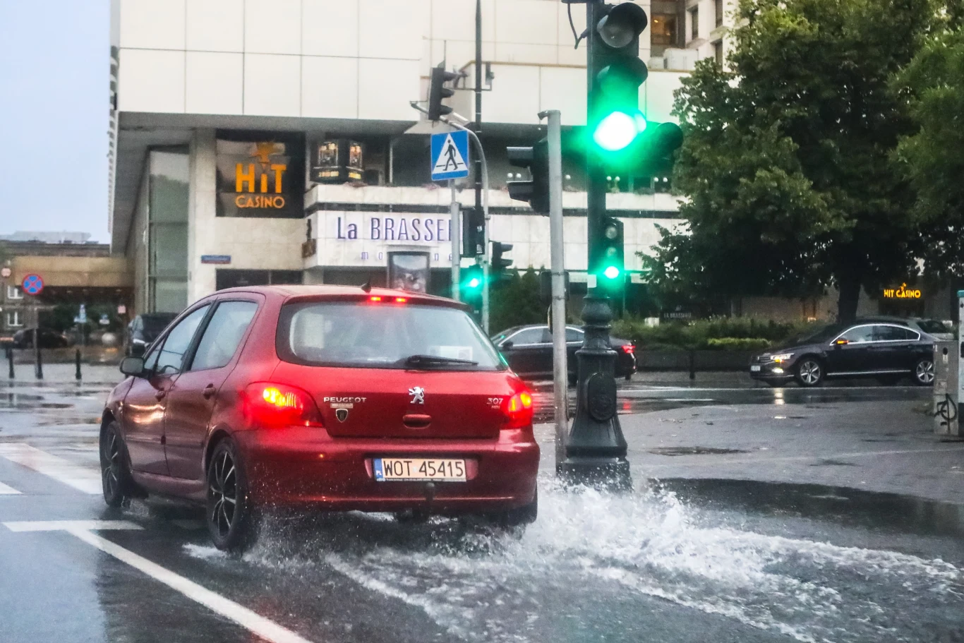 Co grozi za ochlapanie pieszego wodą z kałuży? Czerwony samochód na mokrej jezdni przejeżdża przez kałużę, wzbijając wodę; widoczne budynki i reklamy.