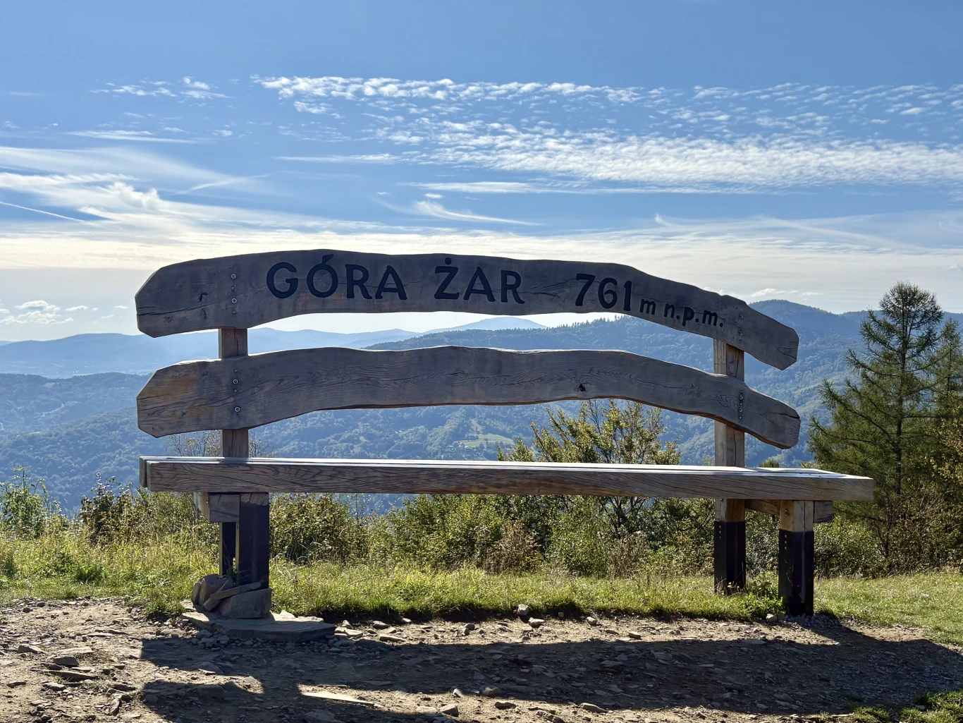 Drewniana ławka z napisem „Góra Żar 761 m n.p.m.” ustawiona na szczycie, z widokiem na malownicze, górskie pasma w tle.