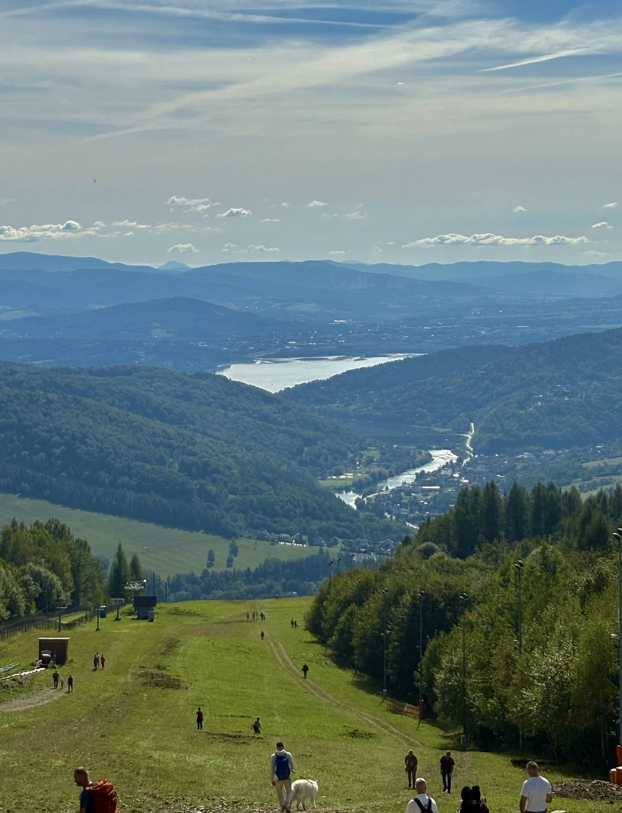 Panorama zalesionych wzgórz z rozległą polaną po środku, przez którą spacerują ludzie, w tle widoczny duży zbiornik wodny oraz góry na horyzoncie.