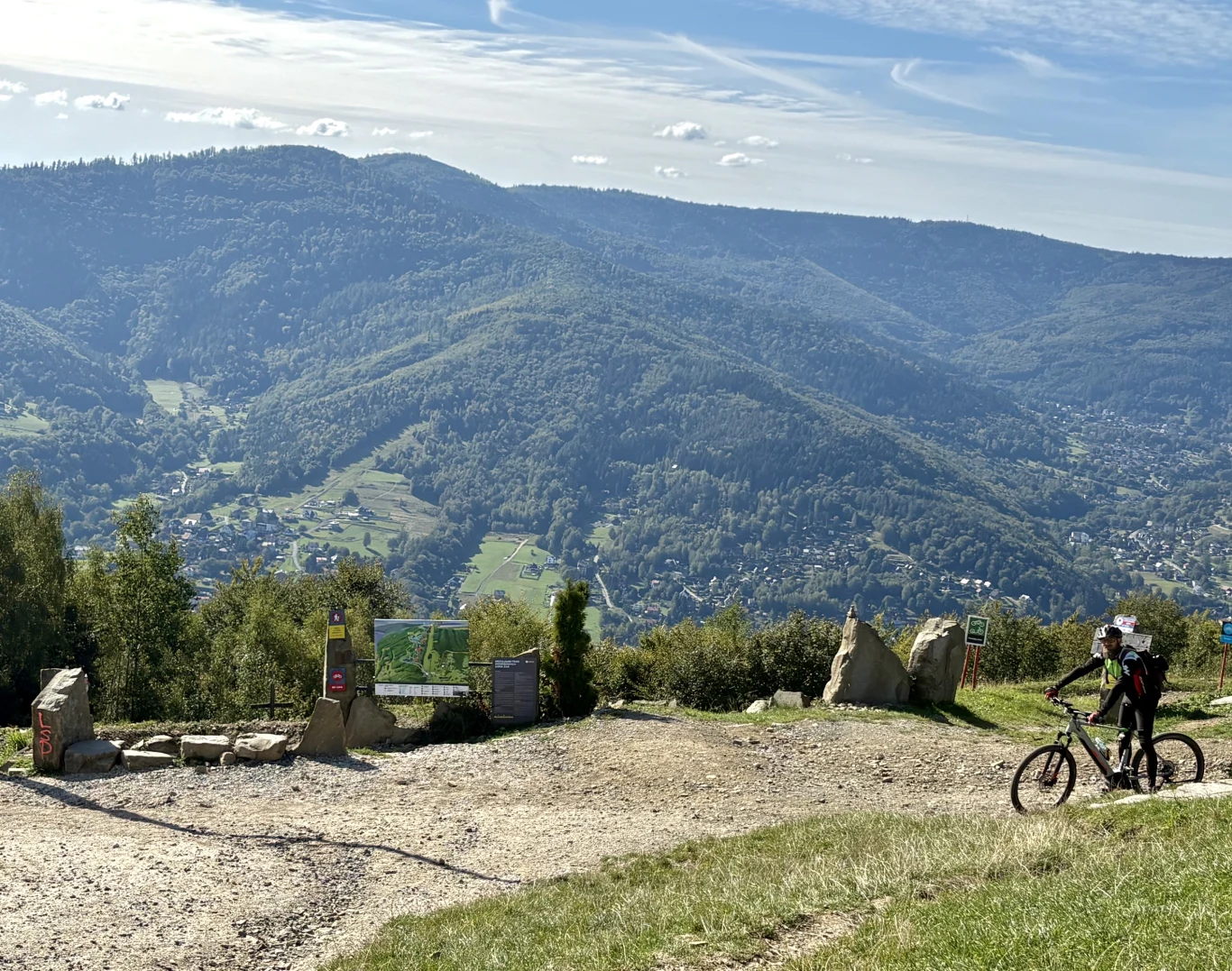 Dwóch rowerzystów w kaskach stoi z rowerami górskimi na szczycie wzgórza, w tle rozciąga się panorama zielonych lasów i gór oraz widoczne są zabudowania miejscowości w dolinie.