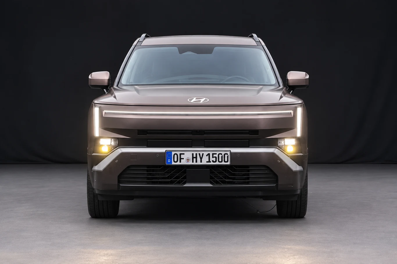 Nowy Tucson wygląda znacznie bardziej kanciasto od poprzednika. Po bokach przodu widać pionowe światła do jazdy dziennej. Brązowy SUV marki Hyundai o nowoczesnym, geometrycznym kształcie z charakterystycznym, poziomym pasem świateł LED i niemiecką tablicą rejestracyjną, sfotografowany na tle ciemnego, jednolitego tła.