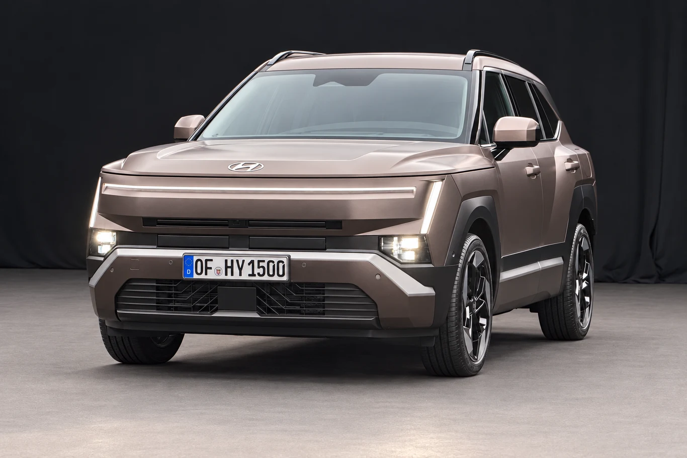 Oświetlenie przednie tworzą pozioma listwa, pionowe światła dzienne i reflektory umieszczone niżej. Brązowy SUV Hyundai z nowoczesnym, geometrycznym designem nadwozia, charakterystycznymi, poziomymi reflektorami LED oraz czarnymi, pięcioramiennymi felgami, ustawiony na szarym podłożu na tle ciemnej kurtyny.