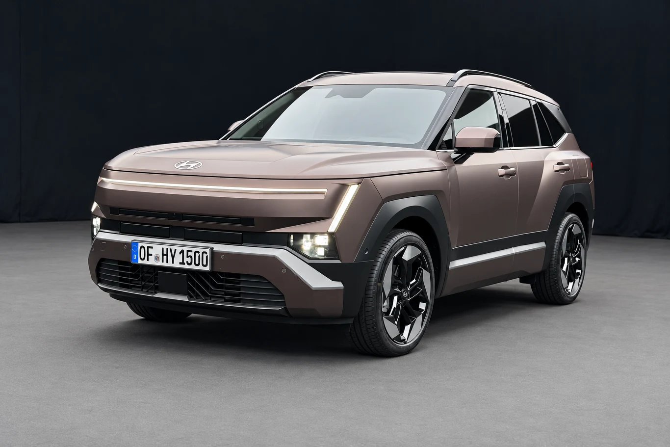 Nowy Hyundai Tucson ma cienką listwę świetlną biegnącą przez cały przód. Właściwe reflektory umieszczono niżej. Nowy Hyundai Tucson, brązowy SUV z cienką listwą świetlną i geometrycznymi liniami, na szarym tle.