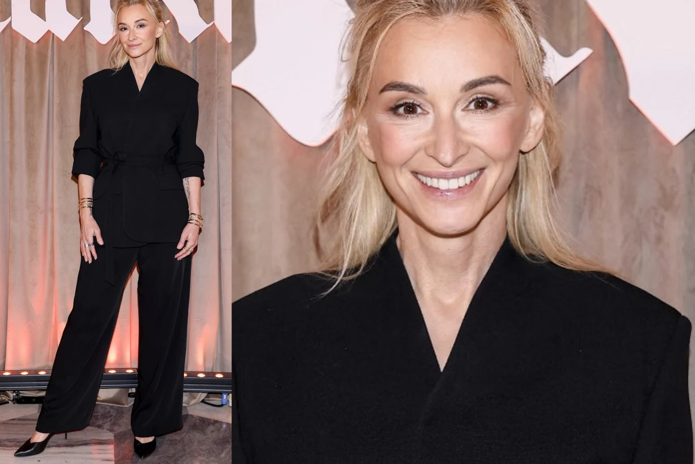 Martyna Wojciechowska na premierze "Lalki" Uśmiechnięta kobieta w eleganckim, czarnym garniturze z szerokimi spodniami, stojąca na tle beżowych zasłon i jasnych elementów scenografii.