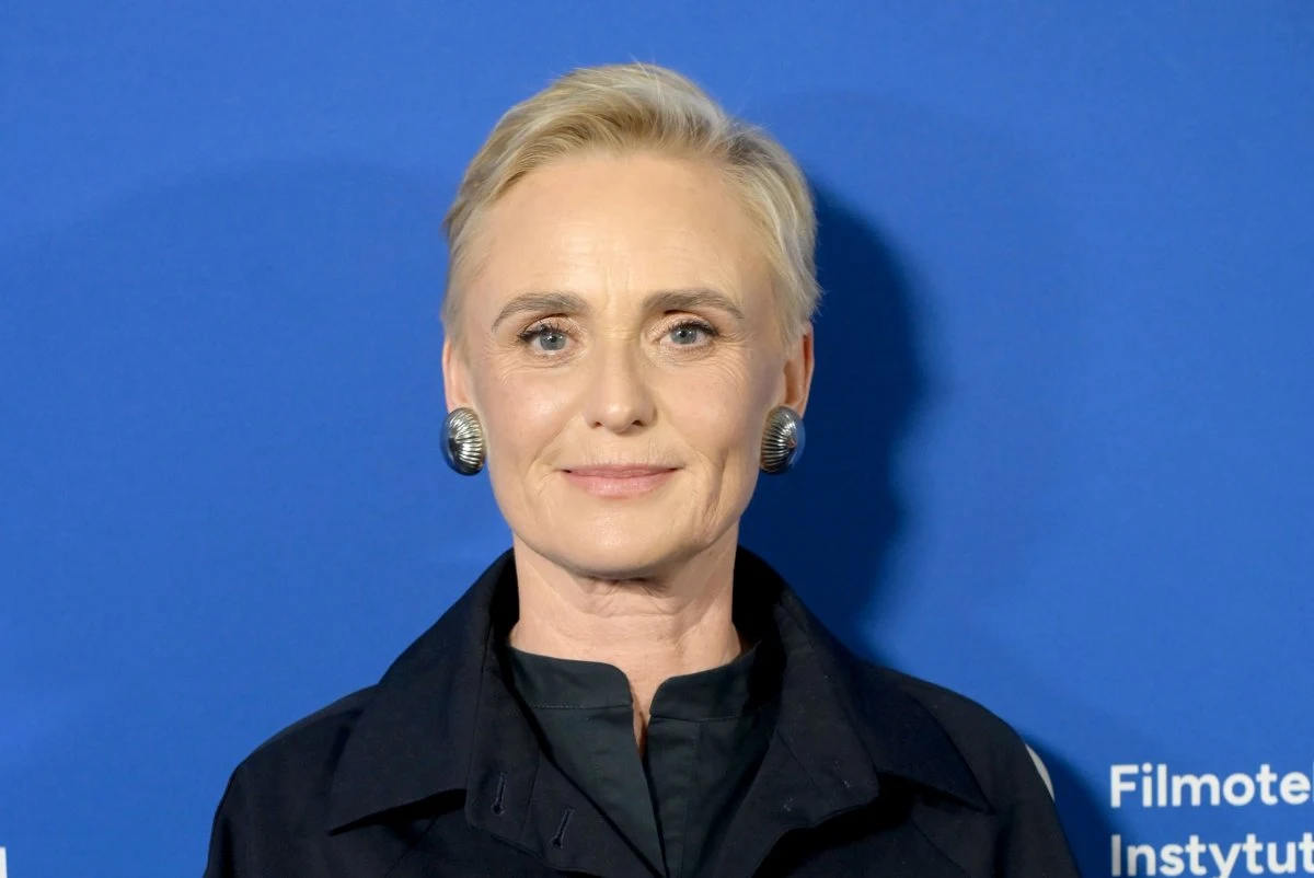 Kinga Preis Kobieta o krótkich blond włosach, w ciemnej marynarce i ozdobnych kolczykach, stoi na tle niebieskiej ścianki z białym tekstem.