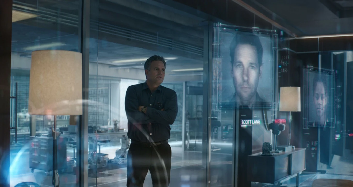 Kadr z filmu "Avengers: Koniec gry" Mężczyzna w ciemnej koszuli stoi w nowoczesnym pomieszczeniu otoczonym szklanymi ścianami, patrząc na duży hologram z wizerunkiem twarzy i podpisem 'Scott Lang', wokół widoczne są lampy oraz inne holograficzne ekrany.