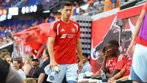 Piłkarz w czerwonym stroju sportowym przechodzi obok ławki rezerwowych podczas meczu piłki nożnej, wokół niego znajdują się inni zawodnicy oraz sztab trenerski, w tle widoczne fragmenty stadionu oraz czerwona grafika na ścianie.