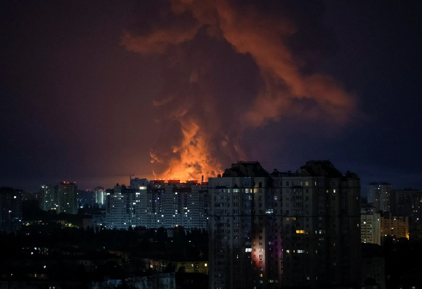 Nocna panorama Kijowa z dużymi płomieniami i kłębiącym się dymem w oddali za wysokimi budynkami mieszkalnymi.