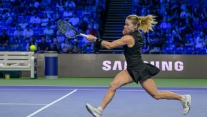 Zawodniczka w czarnej sportowej sukience dynamicznie odbija piłkę tenisową podczas meczu na niebieskim korcie, wykonując skok z rakietą skierowaną do przodu.