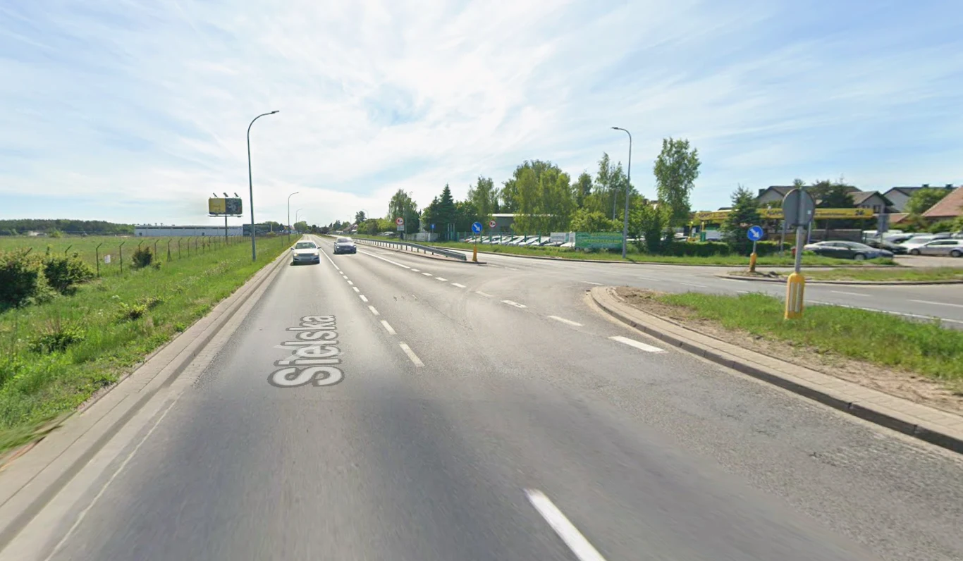 Ul. Sielska w Olsztynie zostanie rozbudowana. Powstanie droga dla pieszych i rowerzystów. Ul. Sielska w Olsztynie, dwupasmowa jezdnia, skrzyżowanie, auta, parking, zabudowania, drzewa, jasne niebo.