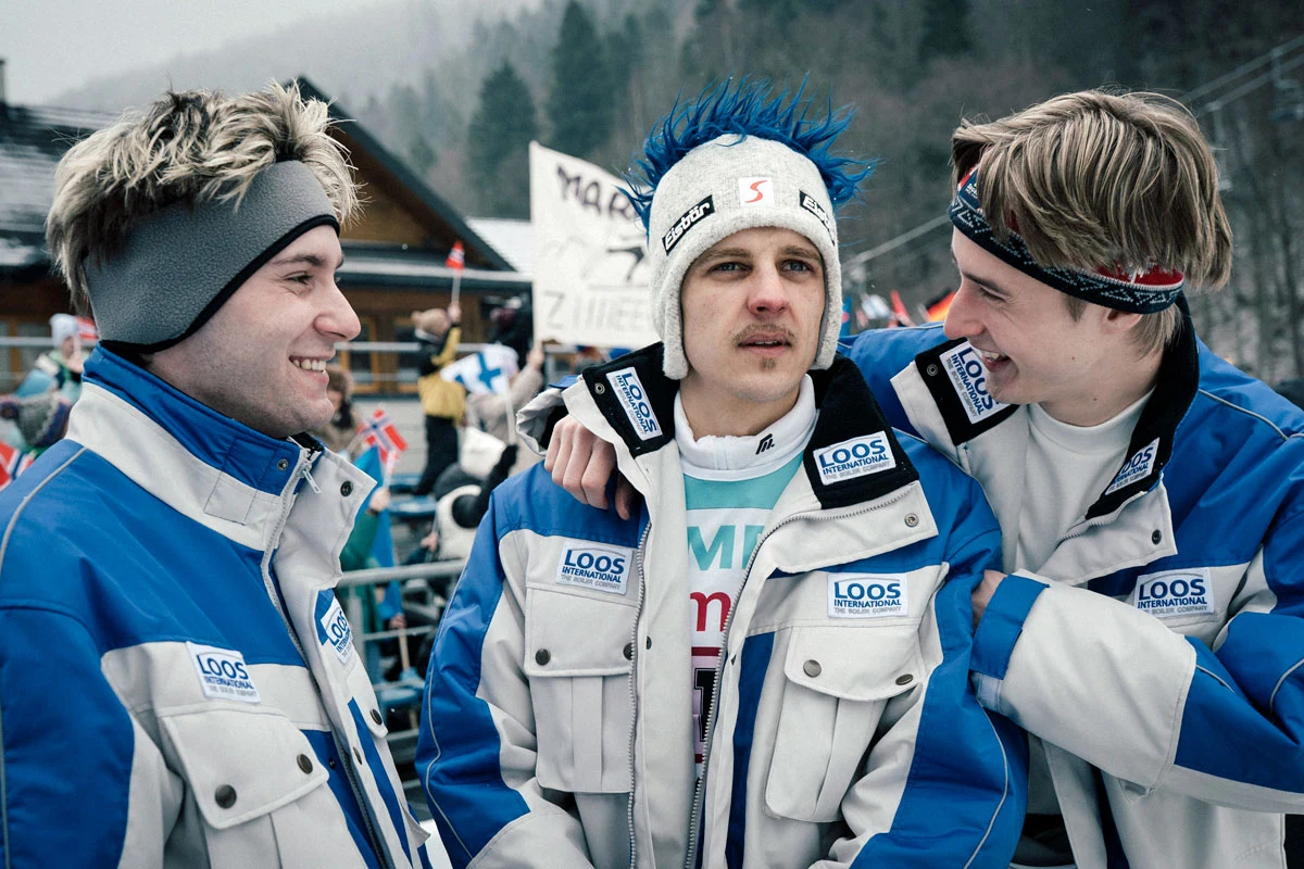 Bartłomiej Deklewa (w środku) jako Adam Małysz Bartłomiej Deklewa jako Adam Małysz w niebiesko-białej kurtce sportowej z dwoma młodymi mężczyznami.