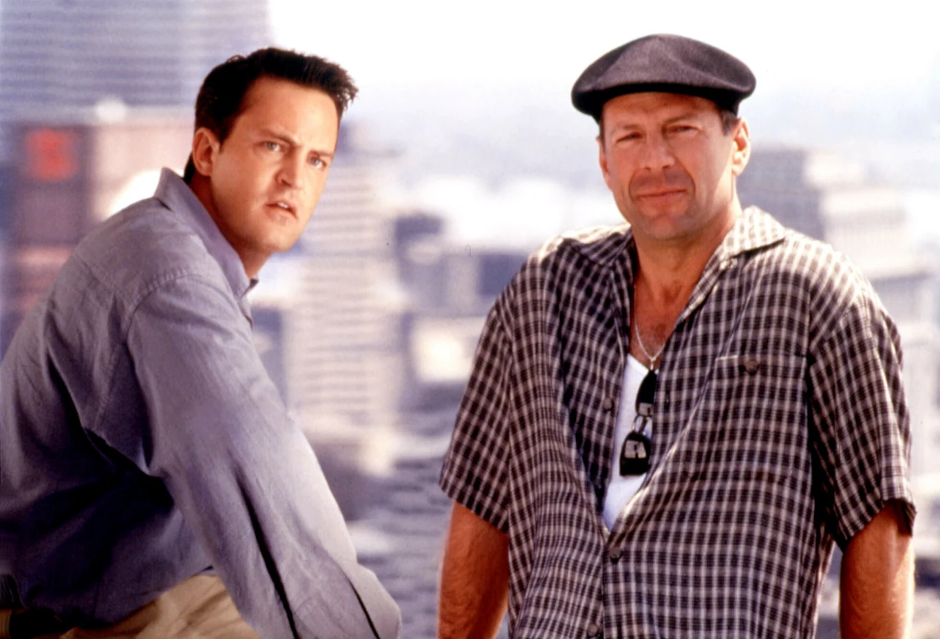 Matthew Perry i Bruce Willis w filmie "Jak ugryźć 10 milionów"? Dwóch mężczyzn stoi na tle panoramy miasta; jeden ubrany w jasnoniebieską koszulę, drugi w koszulę w kratę i czarny beret.