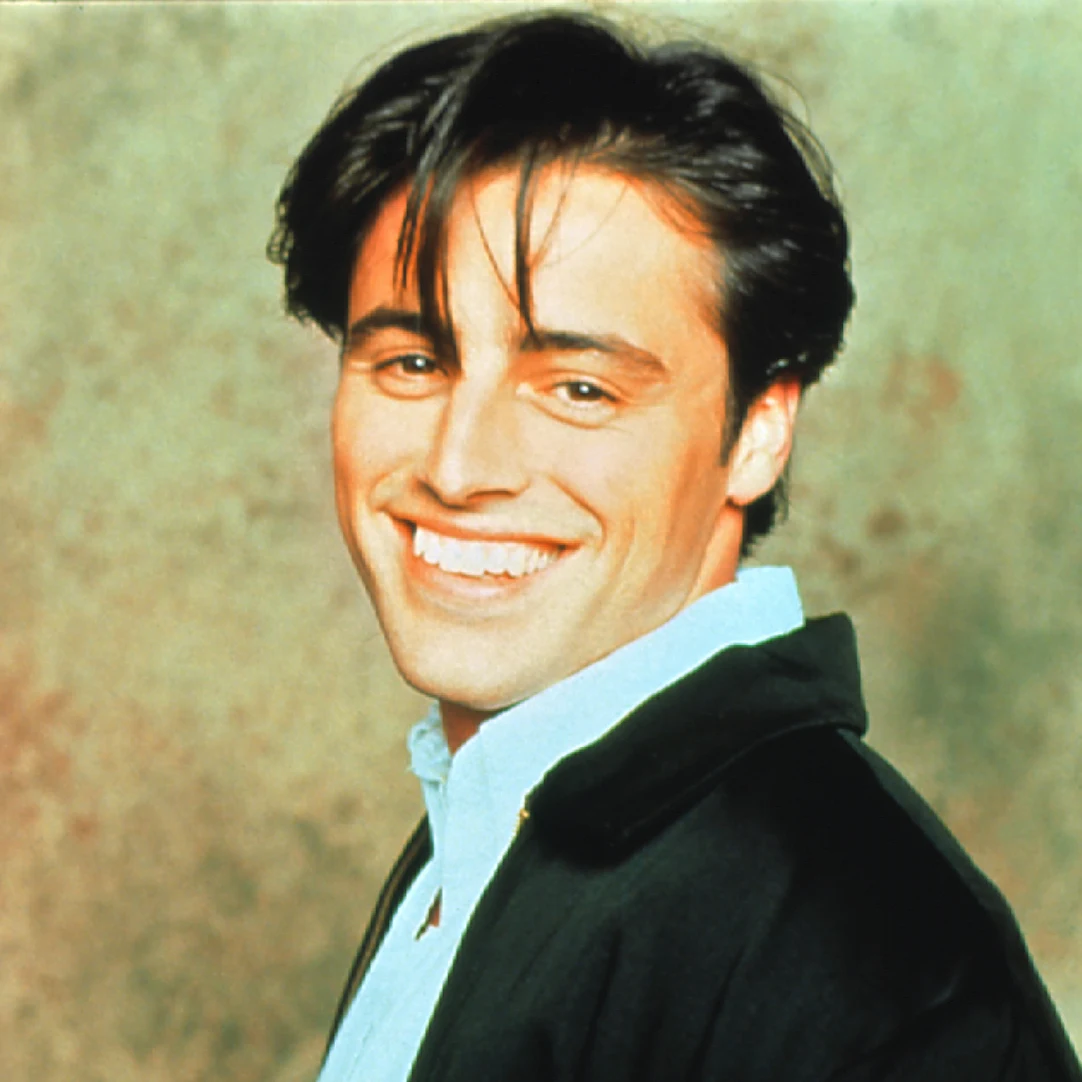 Matt LeBlanc w serialu "Przyjaciele" Uśmiechnięty mężczyzna o ciemnych włosach i jasnej cerze ubrany w białą koszulę i ciemną marynarkę, patrzący w stronę obiektywu.