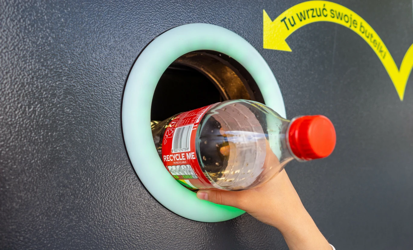 Ręka wkłada plastikową butelkę po napoju do automatu do recyklingu, nad otworem widoczna jest żółta strzałka z napisem zachęcającym do wrzucenia butelki.