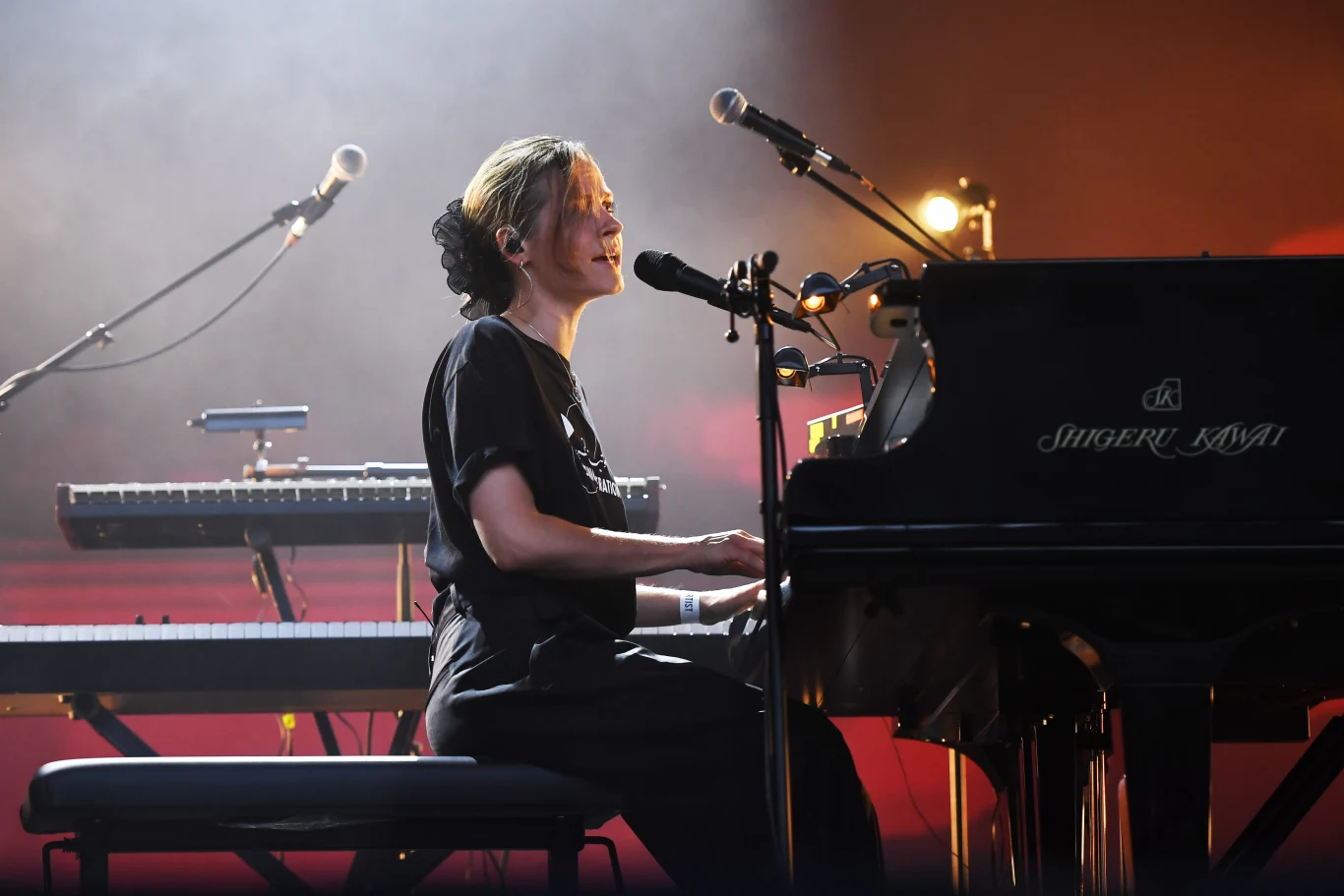Hania Rani podczas występu w Somerset House w Wielkiej Brytanii Kobieta grająca na fortepianie i śpiewająca do mikrofonu na scenie podczas koncertu, w tle klawiatura i światła sceniczne.