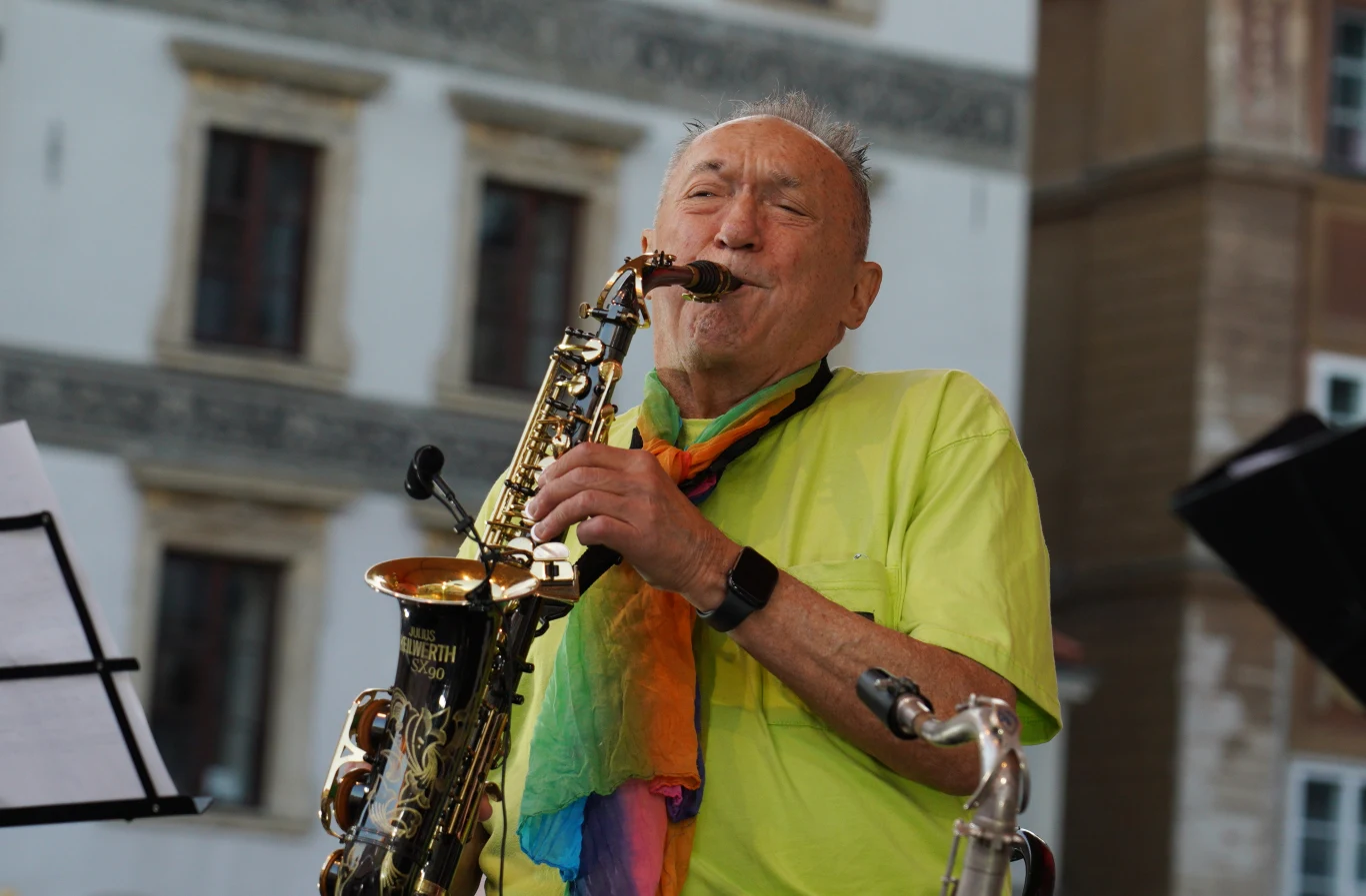 Michal Urbaniak podczas koncertu Jazz na Starówce w 2020 r. Starszy mężczyzna ubrany w jaskrawozieloną koszulkę i kolorową chustę gra na saksofonie, z wyraźnym zaangażowaniem na twarzy; w tle jasny budynek z ozdobnymi oknami.
