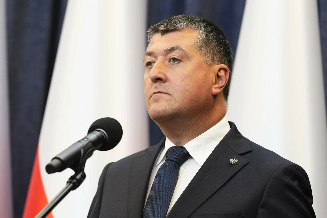 Leszek Skiba jest doradcą prezydenta RP ds. ekonomicznych Leszek Skiba, doradca prezydenta ds. ekonomicznych, przy mikrofonie na tle biało-czerwonych flag.