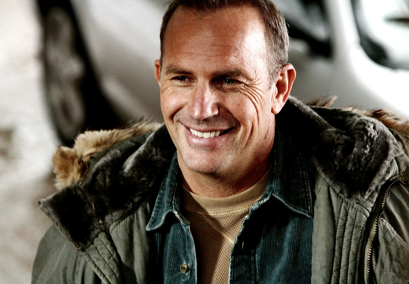 Kevin Costner w filmie "Patrol" Uśmiechnięty mężczyzna w zielonej kurtce z futrzanym kapturem, ubrany warstwowo, patrzący lekko w bok w jasnym otoczeniu.