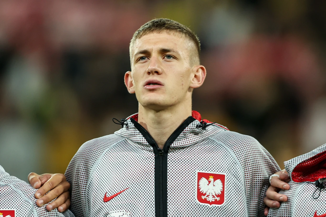 Wiktor Nowak Młody mężczyzna ubrany w sportową bluzę z herbem Polski, stojący z poważnym wyrazem twarzy, trzyma ramię kolegi z drużyny podczas oficjalnej ceremonii.