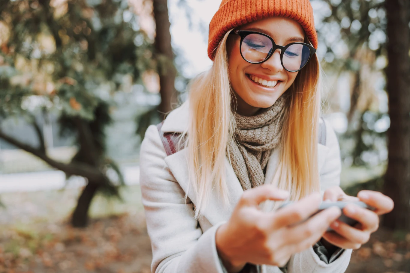 Październik będzie dla nich miesiącem pieniędzy Uśmiechnięta młoda kobieta w okularach i pomarańczowej czapce patrzy na telefon trzymany w dłoniach, ubrana w jasny płaszcz i szalik, na tle drzew.