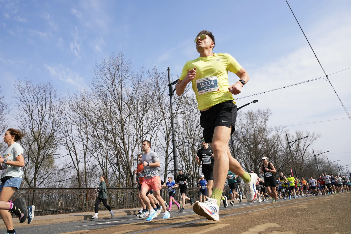 Półmaraton Warszawski Grupa biegaczy na ulicy podczas zawodów, z przodu mężczyzna w żółtej koszulce z numerem startowym, za nim pozostali uczestnicy biegu w sportowych strojach.