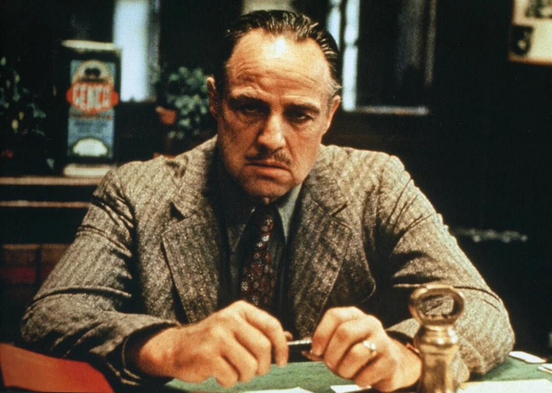 Mężczyzna w szarym garniturze siedzący przy biurku, o poważnym wyrazie twarzy, z dłońmi opartymi o blat i skupionym spojrzeniem skierowanym do przodu.