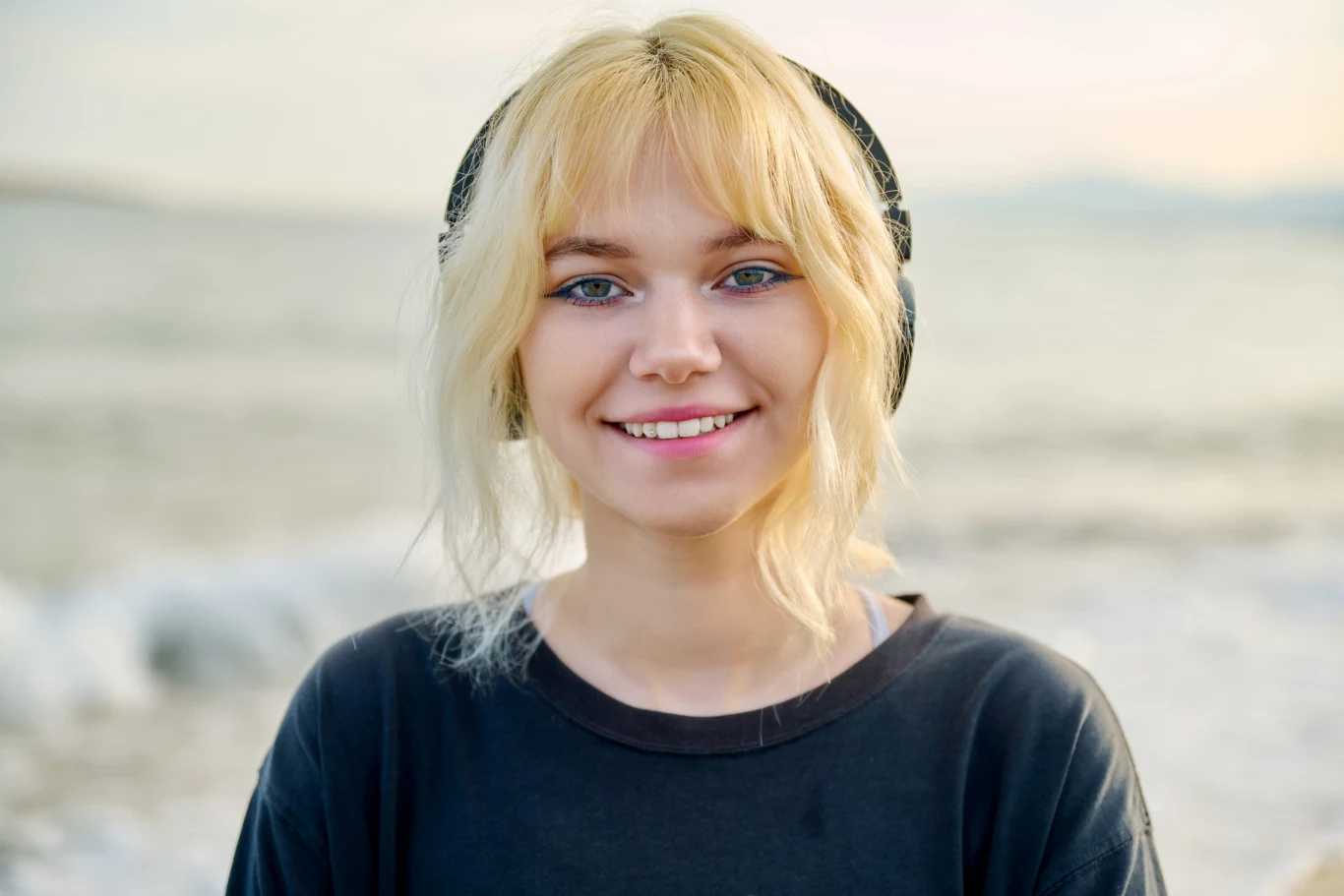 Młoda kobieta z blond włosami i słuchawkami na uszach uśmiecha się, stojąc na plaży przy brzegu morza.